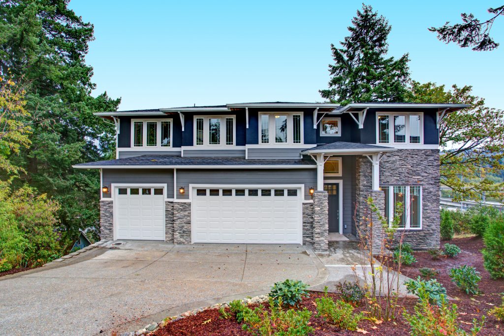 Blue luxury home with stone veneer siding boasts high porch with stone columns, two garage spaces and concrete driveway.