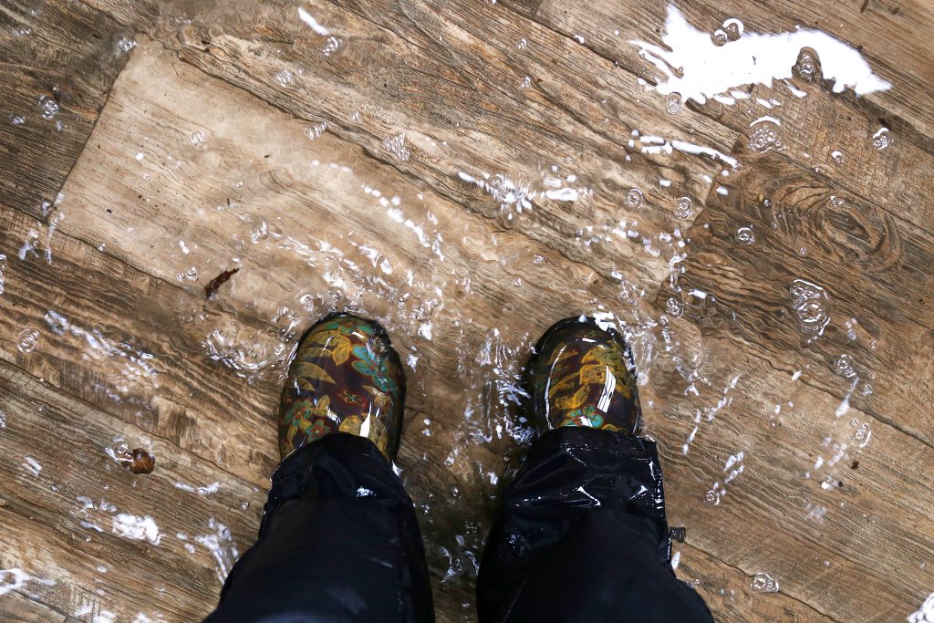 Woman's feet, wearing waterproof rain boots standing in a flooded house with vinyl wood floors, how to spot water damage on wood floor